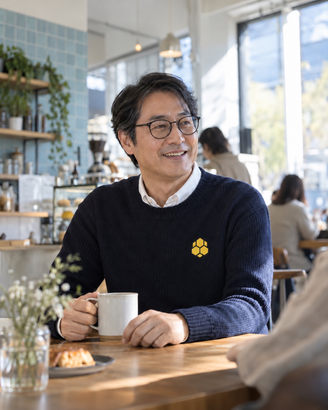 Man in glasses and a navy sweater greeting someone in a sunlit café.